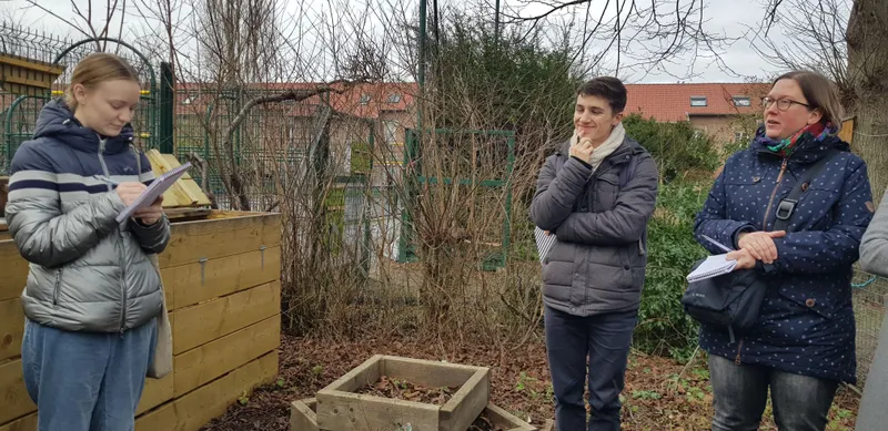 Ein Foto von einer Person, die auf einer Wiese vor einem Baum steht und ein paar Hosen in der Hand hält. Im Hintergrund ist ein Bienenstock und ein Stück Wal...