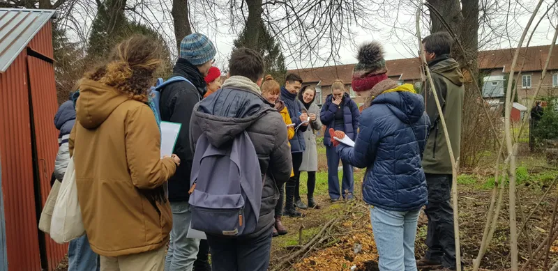 Eine Gruppe von Menschen entspannt sich unter einem Baum und genießt die Sonne. Sie tragen Jeans und Jacken und sind umgeben von Bäumen und Pflanzen. Der Him...
