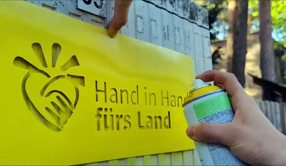 Ein Handgelenk mit gelbem Nagellack hält einen Spraydosen-Kopf in die Luft, der auf einem grasbewachsenen Boden steht. Die Dose zeigt ein Logo mit der Aufsch...