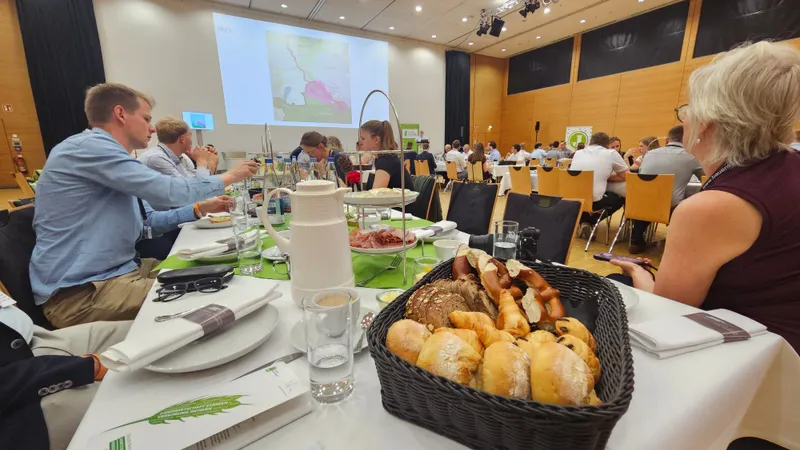 Brötchen im Vordergrund, dahinter ein großer Saal mit vielen Junglandwirt:innen.
