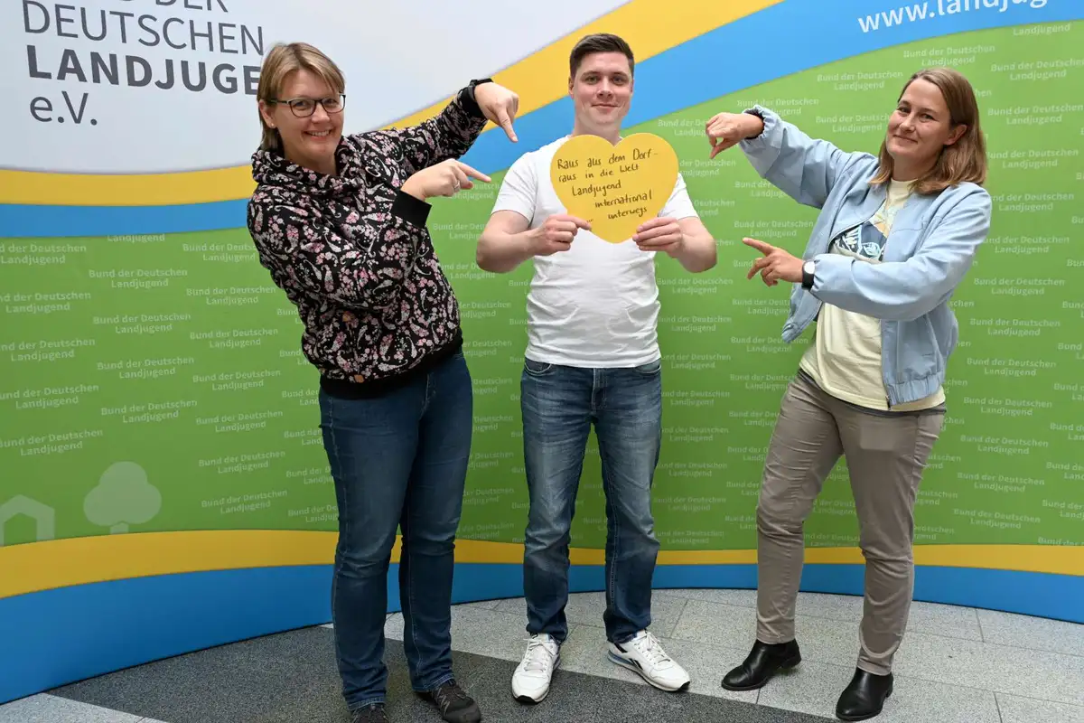 Drei lächelnde Menschen halten ein herzförmiges Schild mit der Aufschrift: Raus aus dem Dorf, Rein in die Welt