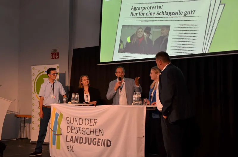 Ein Sprecher hält während einer Veranstaltung eine Rede vor einem Publikum. Er trägt einen Anzug und spricht in ein Mikrofon. Im Hintergrund sind Präsentatio...