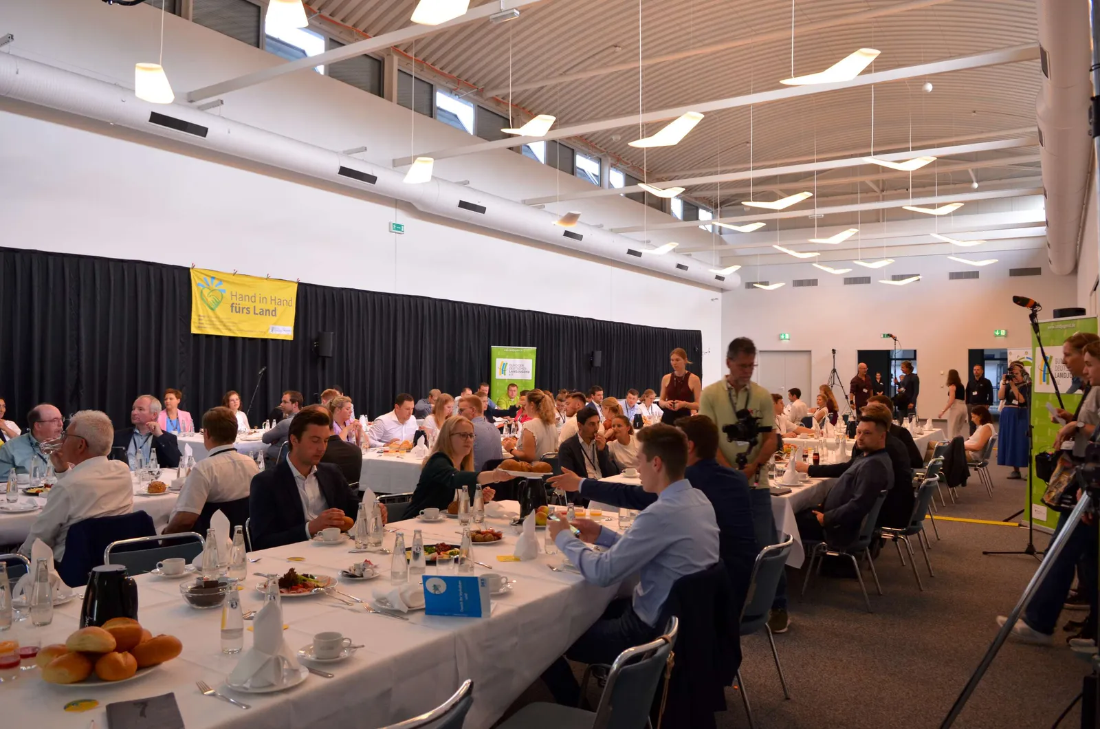 Ein Konferenzraum mit einem großen Tisch, Stühlen und Geschirr. Auf dem Tisch sitzen Menschen in Anzügen bei einem Event. Der Raum wird für eine Konferenz od...