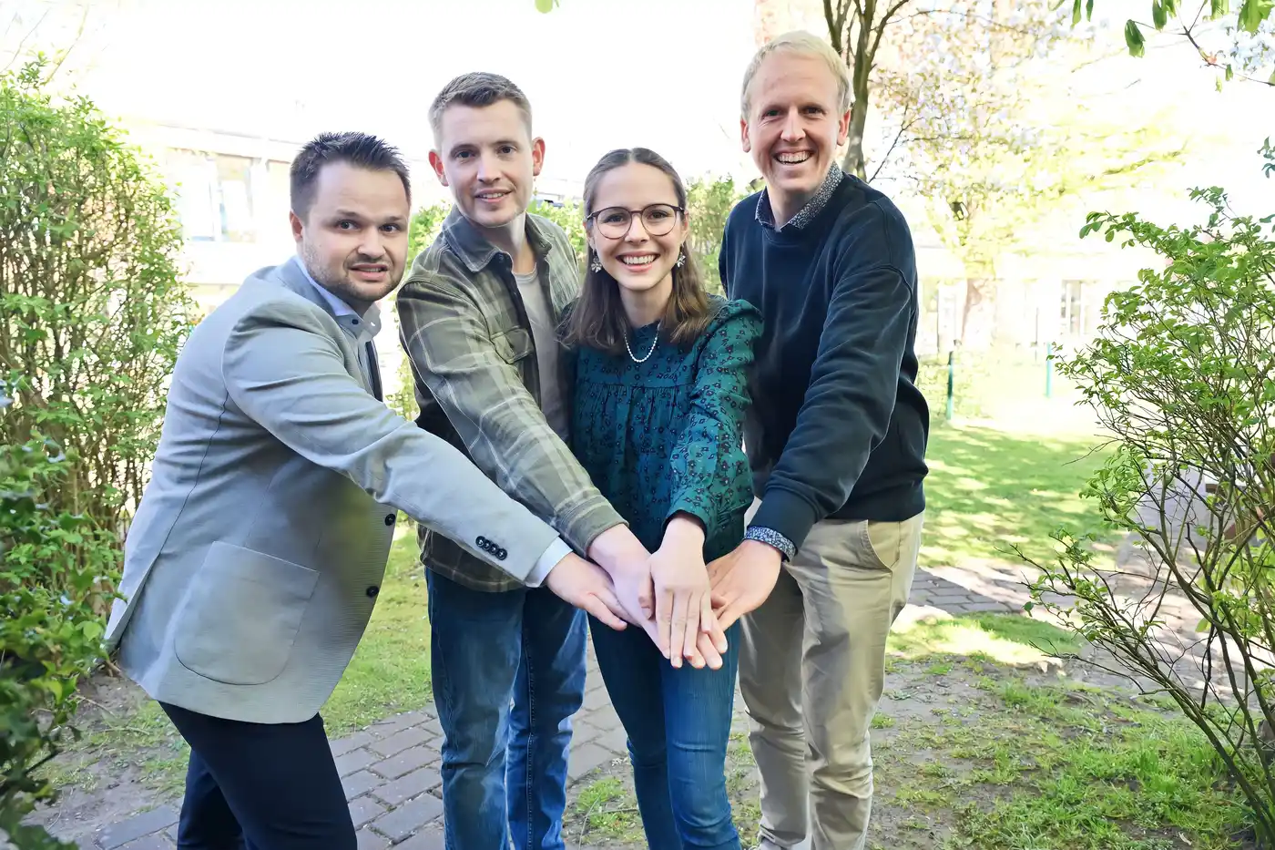 Drei junge Männer und eine junge Frau schauen dich direkt, neugierig und offen an. Jeder hält einen Arm noch vorn und ihre Hände liegen übereinander.