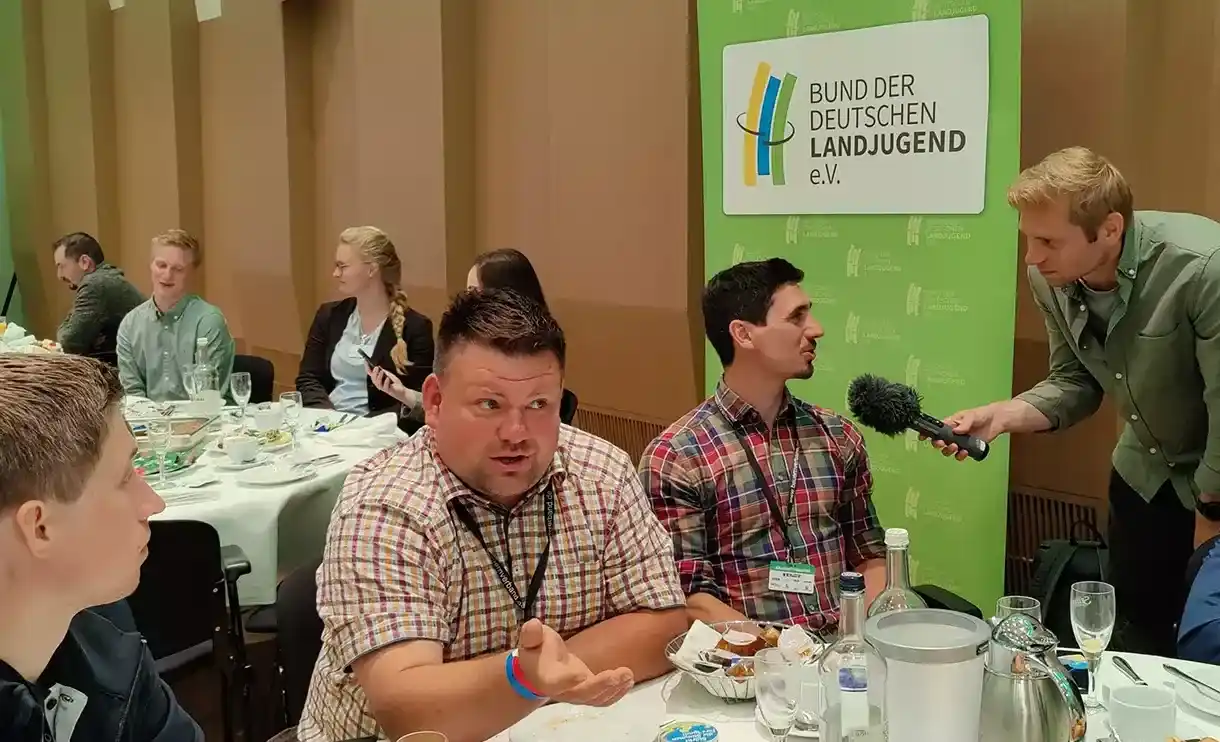 Ein junger Landwirt steht an einem Mikrofon auf einem Tisch während einer Veranstaltung. Er teilt seine Erfahrungen mit anderen Teilnehmern und präsentiert s...
