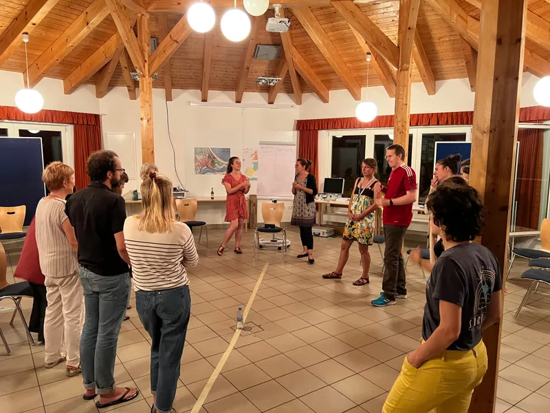 Martina spielt Schlagzeug auf einem Holzstuhl bei einem lustigen Event. Sie trägt eine Hose und genießt die Musik.
