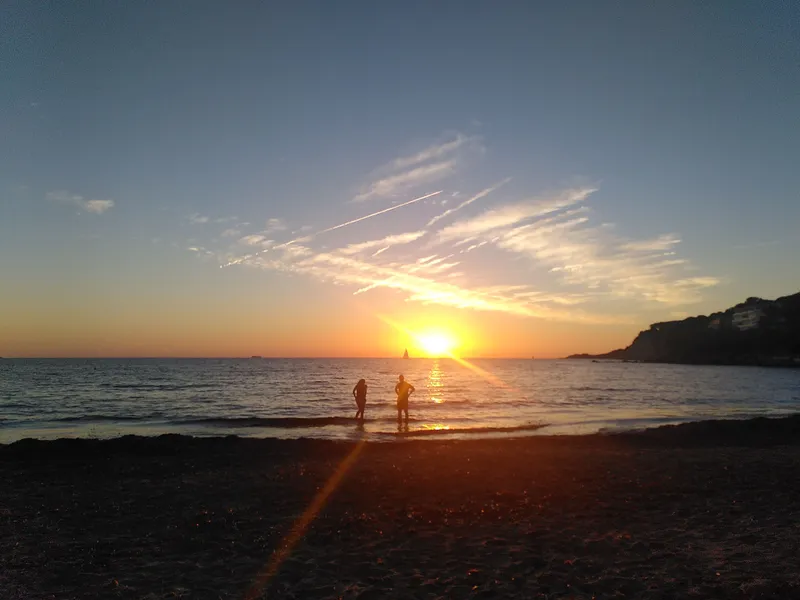 Janina Sarsky am Strand bei Sonnenuntergang. Der Himmel ist in warmen Farben des Abendhimmels gefärbt, während die Sonne hinter dem Horizont versinkt und ein...