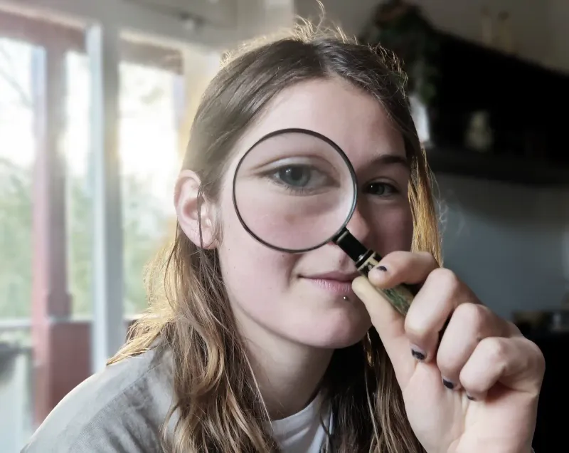 Eine junge Frau mit Brille betrachtet ihr Auge durch eine Lupe. Ihr Auge ist stark vergrößert, sodass man deutlich Wimpern, Augenbrauen und Hautstruktur sehe...