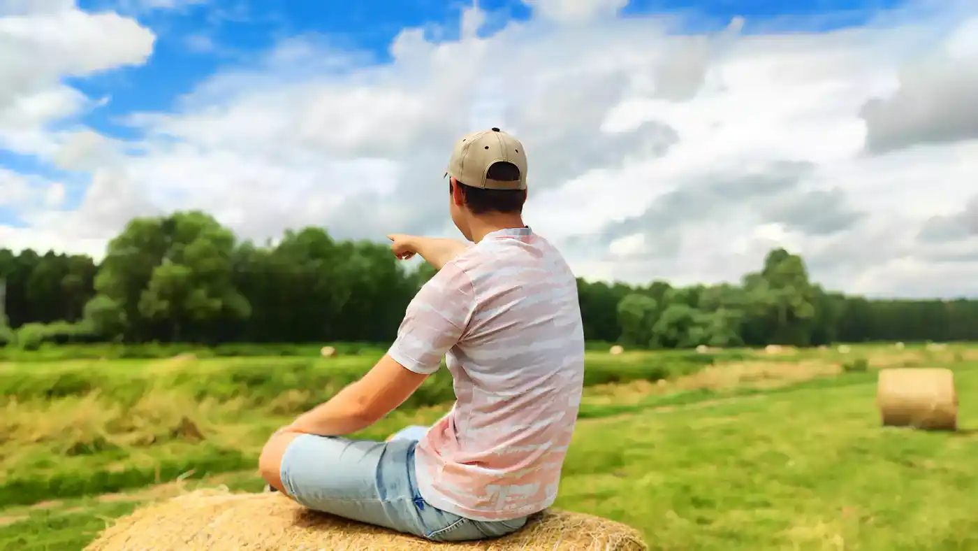 Junge in einem Baseballcap lächelt fröhlich auf einem Feld voller Gras und Strohballen. Der Himmel ist klar und sonnig, mit ein paar weißen Wolken. Er genieß...