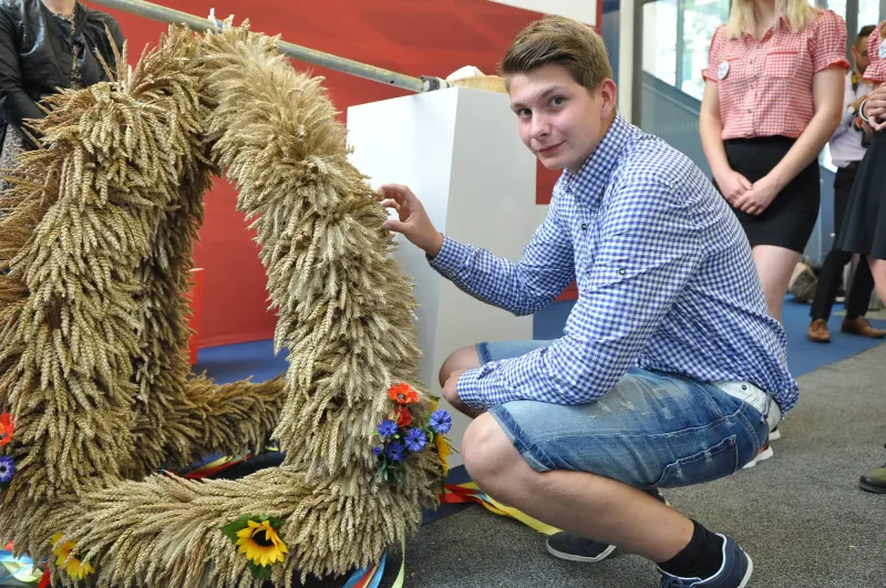 Ein junger Mann mit einem breiten Lächeln trägt eine gelbe, mit Fell besetzte Erntekrone und einen karierten Rock. Er posiert für ein Event, seine blauen Sch...