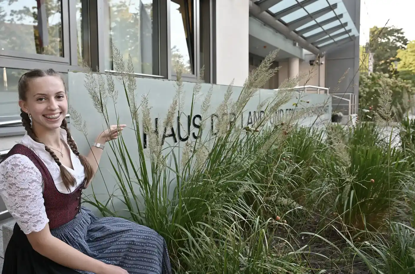 Zwei glückliche Landjugendliche in Tracht stehen lächelnd auf einer grünen Wiese vor einem Schild mit der Aufschrift "Haus der Land- und Ernährungswirtschaft...