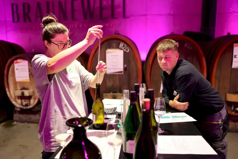 Ein Jungwinzer in modischer Kleidung präsentiert seine Weine bei einem Berufswettbewerb im Weinkeller. Auf dem Tisch stehen Flaschen und Gläser mit Wein. Die...
