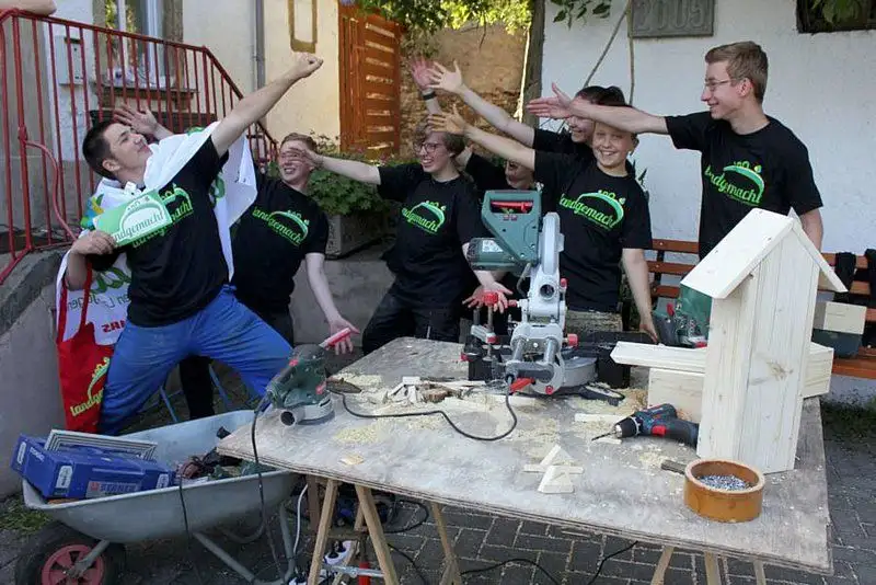 Eine Gruppe von Jugendlichen sitzt an einem Tisch vor einem Fenster bei einer Freizeitaktivität. Auf dem Tisch steht eine Pflanze. Die Teilnehmer tragen T-Sh...