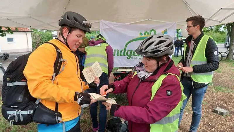 Ein lächelnder Radfahrer trägt einen grünen Fahrradhelm, eine Jeans und eine Jacke. Im Vordergrund sind Fahrradreifen und weitere Radutensilien zu sehen.  
