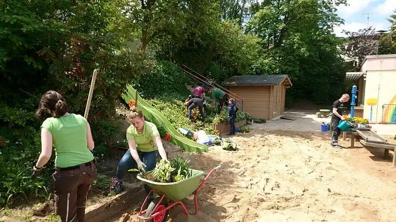 Zwei Personen genießen einen entspannten Tag im Garten. Ein Mann schiebt eine Schubkarre mit Reifen und Gartengeräten auf grünem Rasen neben einem Baum und e...