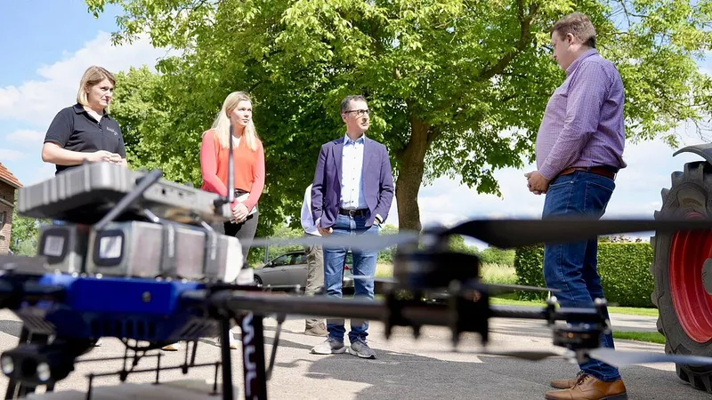 Musiker spielt auf einer Trommel im Freien, umgeben von Bäumen und Grünflächen. Im Hintergrund ein asphaltierter Weg und ein Auto.