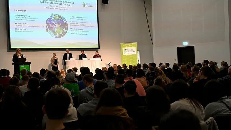 Ein Redner in Anzug hält eine Präsentation vor einem Publikum auf einer Bühne. Das Publikum sitzt auf Stühlen und ist gespannt auf die Ausführungen. Im Hinte...