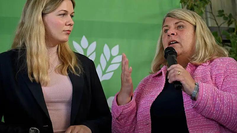 Eine Person spricht in ein Mikrofon während einer Veranstaltung. Sie lächelt und blickt in die Kamera. Die Aufnahme zeigt eine Person in einem sozialen Konte...