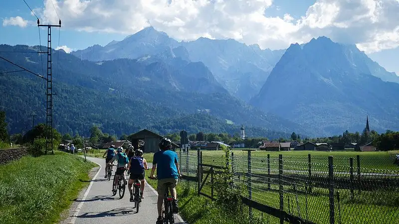 Ein Tandemfahrrad fährt auf einem Weg durch eine malerische Landschaft. Blauer Himmel mit weißen Wolken über schroffen Bergen und üppigem Grün. Die Räder des...