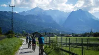 Ein Tandemfahrrad fährt auf einem Weg durch eine malerische Landschaft. Blauer Himmel mit weißen Wolken über schroffen Bergen und üppigem Grün. Die Räder des...