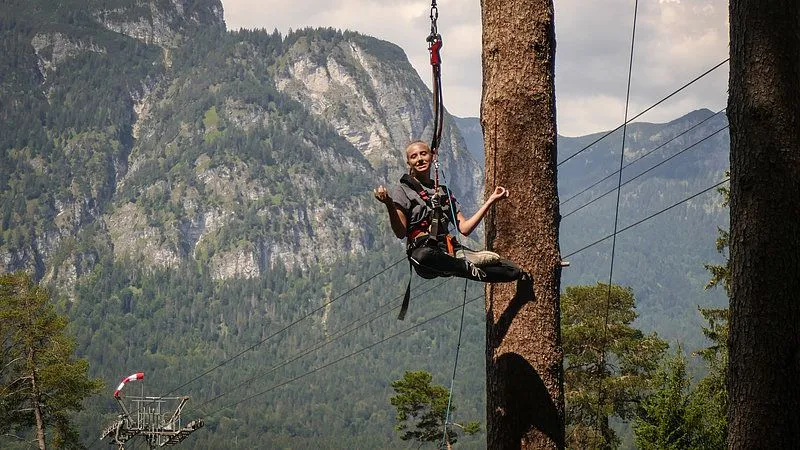 Ein Stuntman klettert an einem Felswand in den Bergen. Der Himmel ist bedeckt mit Wolken und im Hintergrund sind Bäume zu sehen. Der Stuntman trägt Kletterau...