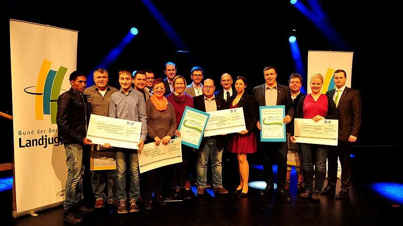 Ein Lächelnder Gewinner der csm_eeg_preis 2017 Auszeichnung auf der Bühne in Jeans, Jacke und Hose. Die Bühne ist mit Mikrofonen und Lautsprechern ausgestatt...